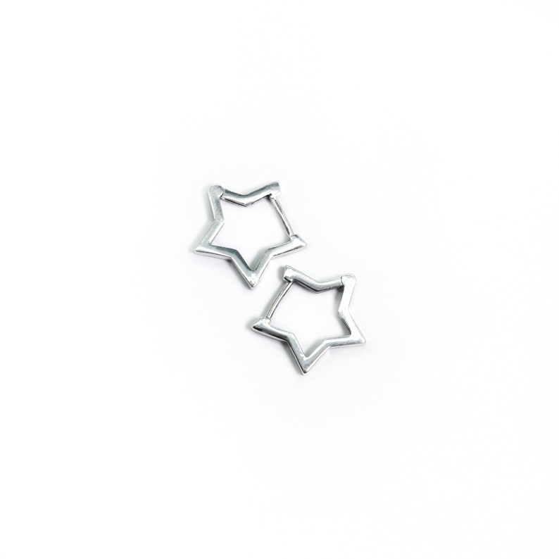 May include: Two silver-colored star-shaped earrings. The earrings are open in the center and have a five-point star design. The earrings are on a white background.