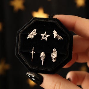以下が含まれることがあります： 黒い八角形のベルベット製ディスプレイに配置された6つの銀色のスタッドピアス。デザインには、蛾、五芒星、コウモリ、短剣、鳥の頭蓋骨、小さな石が埋め込まれた開いた手のひらが含まれています。