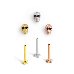 Könnte beinhalten: Ein Set aus drei totenkopfförmigen Körperschmuck-Steckern in Silber-, Gold- und Roségoldtönen, jeweils mit schwarzen Augenhöhlen. Das Set enthält drei passende Metallstifte mit sternförmigen flachen Rückseiten zur sicheren Befestigung.