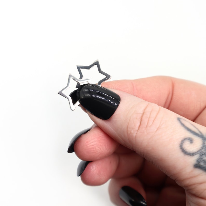 May include: A pair of silver star-shaped earrings. The earrings are held in a hand with black nail polish against a white background. The earrings are small and delicate, with a simple, open design.