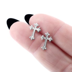 Könnte beinhalten: Zwei silberfarbene Kreuz-Ohrstecker. Jeder Ohrring hat ein detailliertes, verziertes Kreuzdesign mit dekorativen Verzierungen. Die Ohrringe sind klein und filigran, geeignet für den täglichen Gebrauch.