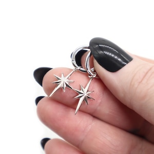 Pode incluir: Um par de brincos de argola prateados com pingentes de estrela pendentes. Cada pingente tem oito raios pontiagudos que irradiam de um ponto central, criando um design celestial simétrico com um acabamento polido e brilhante.