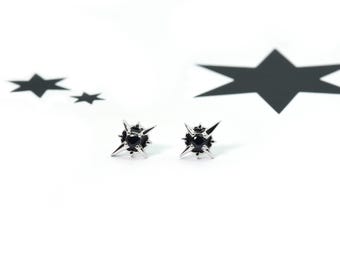 May include: A pair of silver stud earrings featuring a central round black stone surrounded by four pointed starburst rays. The earrings are positioned in front of a white background with two black star shapes printed on the surface.