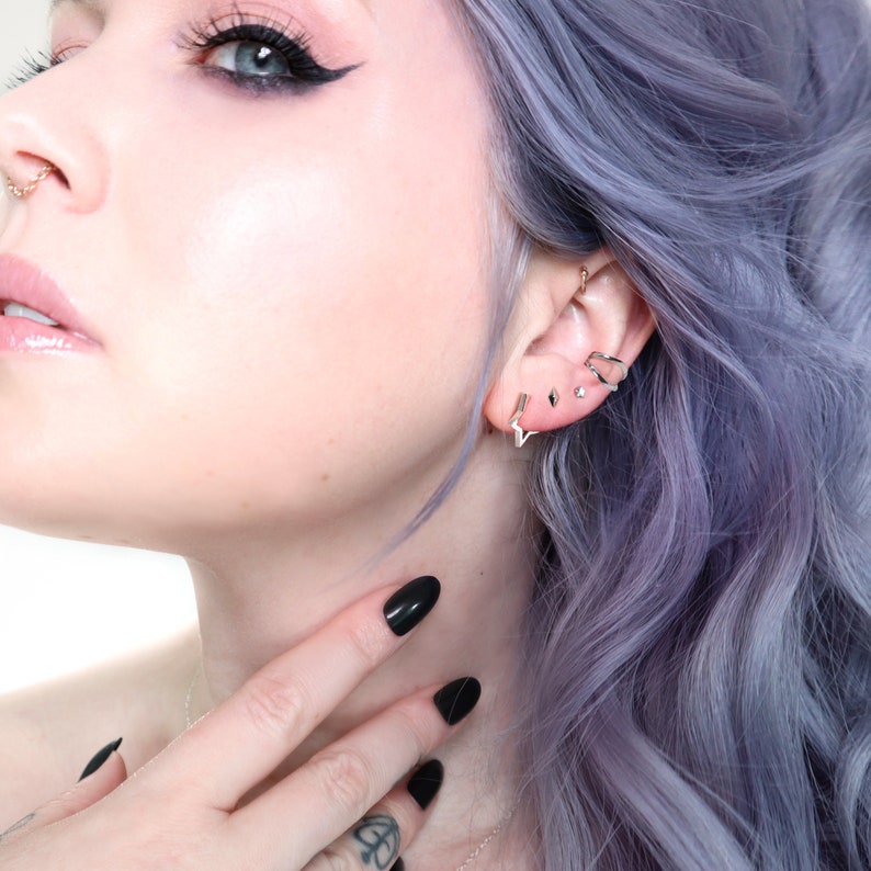 May include: A close-up of a person's face and ear adorned with multiple silver earrings. The earrings include a star-shaped stud, a small diamond-shaped stud, and a curved ear cuff. The person has black nail polish and blue-purple hair.