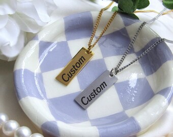 May include: Two gold and silver necklaces with rectangular name tags that say "Custom" on a white and purple checkered surface.