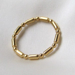 Puede incluir: Un anillo dorado con un diseño de cuentas. El anillo presenta cuentas cilíndricas y esféricas alternas, creando una apariencia texturizada y elegante. El anillo está sobre un fondo blanco.