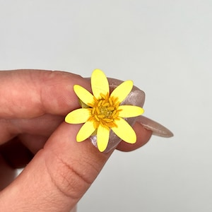 以下が含まれることがあります： 複数枚の花びらと質感のある中心部を持つ、鮮やかな黄色の花のクローズアップ。 花は指の間に挟まれ、繊細なディテールが際立っています。 花びらは鮮やかな黄色で、中心部はより濃い黄色の色合いです。