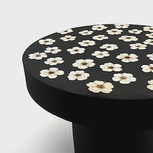 May include: A round black table with a flat top surface decorated with a repeating pattern of pressed white flowers. The table features a solid cylindrical base and a smooth matte finish, showcasing the delicate petals of the dried blossoms.