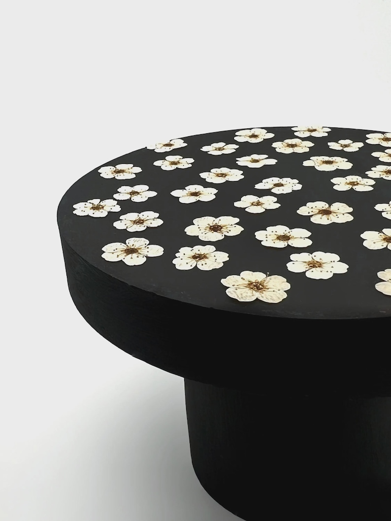 May include: A round black table with a flat top surface decorated with a repeating pattern of pressed white flowers. The table features a solid cylindrical base and a smooth matte finish, showcasing the delicate petals of the dried blossoms.