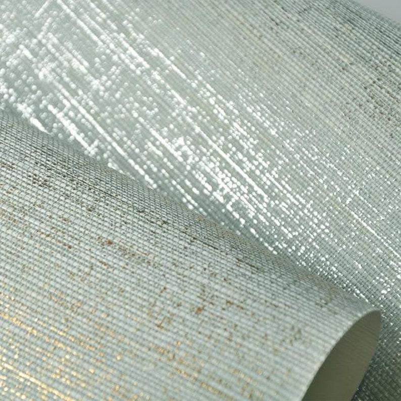 Op de afbeelding: Twee rollen textuurbehang, een met een zilveren glans en de andere met een subtiele gouden glans. Beide rollen hebben een geweven, linnenachtige textuur.