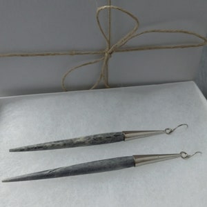 May include: A pair of long, silver earrings with a gray, textured, wooden-like material. The earrings have a pointed tip and a silver hook.