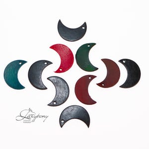 Puede incluir: Una colección de diez colgantes con forma de media luna en varios colores, incluyendo negro, rojo, verde y marrón oscuro. Cada colgante tiene un pequeño orificio circular en la parte superior para colgarlo. El texto Lazypony aparece impreso abajo.