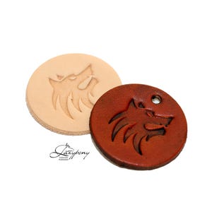 Peut inclure: Deux étiquettes circulaires en cuir arborant un motif de tête de loup. Une étiquette est beige clair, l'autre est brun rougeâtre foncé avec un petit trou percé près du bord supérieur. Le logo Lazypony est imprimé en noir en bas à gauche.