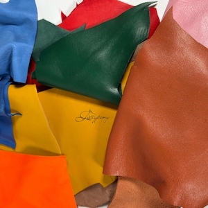 May include: A collection of assorted leather scraps in various colours including blue, red, green, yellow, orange, brown, and pink. The yellow piece features a black logo with the text "The Leather Guy" printed in a cursive script font.