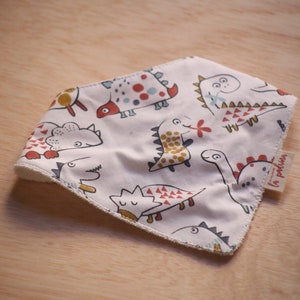 May include: White baby bib with a repeating pattern of colorful cartoon dinosaurs. The bib has a soft, absorbent texture and a tag that reads "la padrima". The bib is designed to catch spills and drool.
