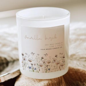 Peut inclure: Une bougie en verre blanc avec une étiquette indiquant "Vanilla Birch". L'étiquette présente des illustrations florales et le texte "Soy Wax Candle 4 oz / 113 g". La bougie est posée sur un tissu beige.