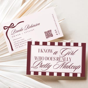 May include: Two business cards with text, one reading "Brooke Robinson, Owner | Stylist" and the other stating "I know a Girl Who Does Really Pretty Makeup." The cards are burgundy and white, with a bow design. A dried palm leaf is in the background.