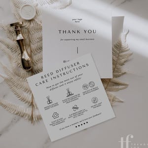 May include: A flat lay featuring a gold-toned diffuser bottle stopper, a white wax seal, and two cards with text. One card reads "THANK YOU" and the other provides reed diffuser care instructions. The background includes white fern leaves.