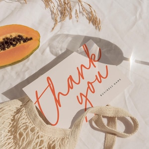 Puede incluir: Imagen de naturaleza muerta con una tarjeta de felicitación con la frase "thank you" en escritura roja. También hay media papaya, un vaso de agua y una bolsa de cuerdas beige. La escena se desarrolla sobre una superficie de lino blanco.