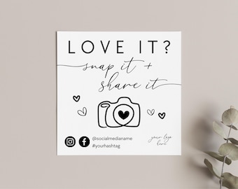 May include: A square white sign with black text reading LOVE IT? snap it + share it. The design features a line-art camera icon with a heart in the center, four small heart outlines, social media icons, and placeholders for a handle and hashtag.