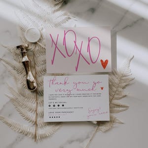 May include: A white greeting card with pink XOXO text and a red heart, paired with a thank you card featuring black and pink text, social media icons, and a five-star graphic. A metallic wax seal stamp rests beside the cards on a white fern leaf.