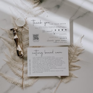 Puede incluir: Una composición plana con dos tarjetas blancas con texto negro, un sello de cera dorado y un sello de lacre dorado. Las tarjetas dicen "Gracias" y "Cuidado de la tabla de cortar". El follaje blanco seco está dispuesto alrededor de las tarjetas.