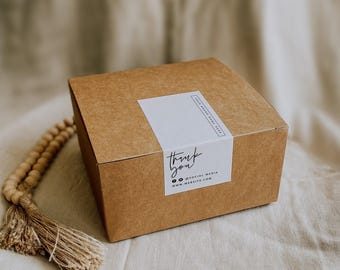 May include: A square brown cardboard box sealed with a white rectangular sticker featuring the text thank you, social media icons, and a website address. A string of light wooden beads with a jute tassel rests beside the box.