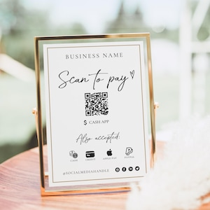 Puede incluir: Un letrero con marco dorado con el texto "BUSINESS NAME" y "Scan to pay". Incluye un código QR, "$ CASH APP" e iconos de métodos de pago aceptados: efectivo, crédito, Apple Pay y PayPal. El letrero está sobre una superficie de madera.