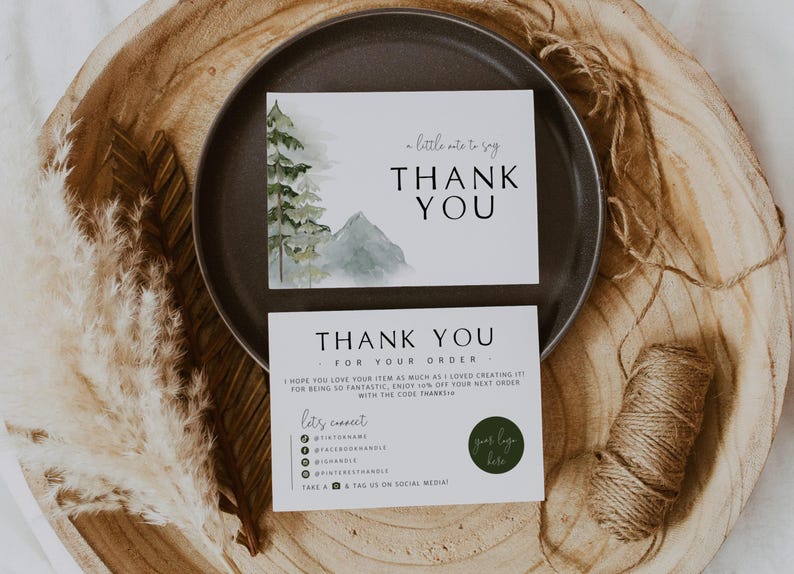 May include: Two white cards with "THANK YOU" printed in black. One card has a watercolor image of a mountain and trees. The other card says "THANK YOU FOR YOUR ORDER" and has social media icons. The cards are on a dark gray plate and a wooden tray.