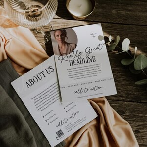 May include: Two white business flyers with the text "Really Great Headline" and "About Us" are displayed on a wooden surface. The flyers feature a photo of a woman and the words "call to action". A glass, candle, and fabric are also visible.