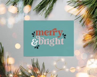 May include: A rectangular greeting card with a teal background featuring the text merry in red and & bright in light pink, accented by small black leaf-like shapes. The design is centered on the card with a clean, bold typography.