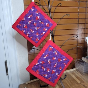Peut inclure: Deux maniques carrées matelassées avec un motif violet de chapeaux rouges et d'os blancs. Les maniques ont une bordure rouge et sont suspendues à un présentoir en métal décoratif.