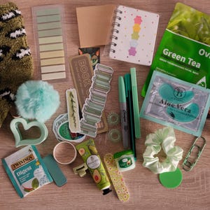 May include: An assortment of self-care items, including a green and white leopard print sock, a small notebook, pens, face masks, and a scrunchie. Other items include stickers, a heart-shaped cookie cutter, and a packet of hot chocolate.