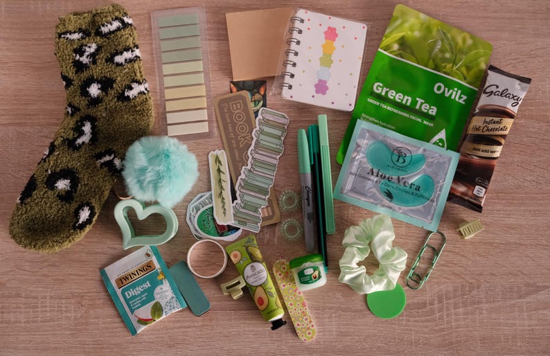 May include: An assortment of self-care items, including a green and white leopard print sock, a small notebook, pens, face masks, and a scrunchie. Other items include stickers, a heart-shaped cookie cutter, and a packet of hot chocolate.