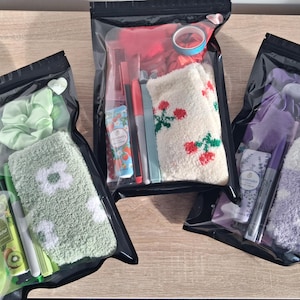May include: Three clear, sealed bags containing various self-care items. One bag has green items, including a scrunchie and socks with a white flower design. Another bag has red items, including socks with red floral designs. The third bag has purple items.
