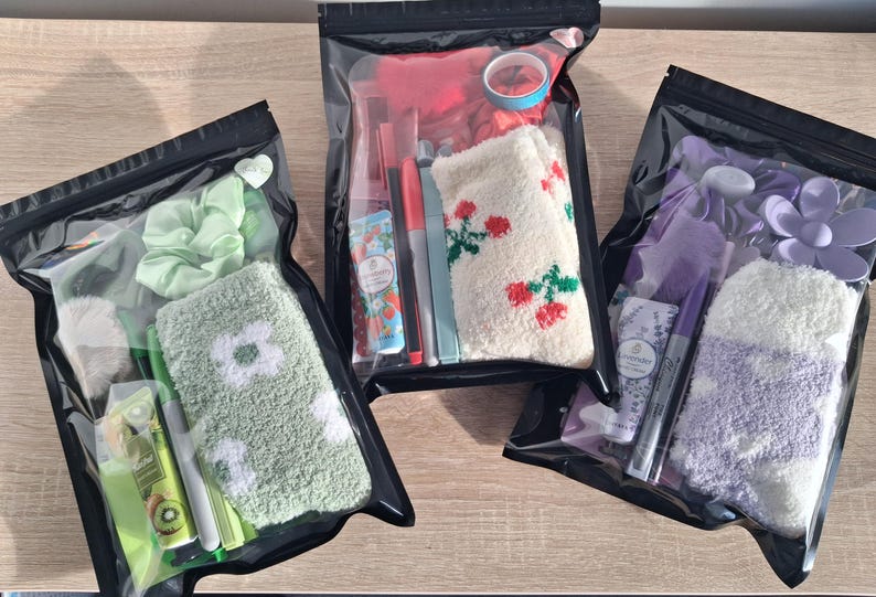 May include: Three clear, sealed bags containing various self-care items. One bag has green items, including a scrunchie and socks with a white flower design. Another bag has red items, including socks with red floral designs. The third bag has purple items.