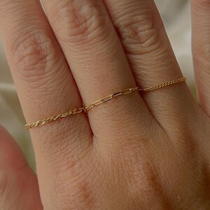 May include: Two gold chain rings on a hand. Both rings are thin and delicate, with one featuring a more intricate chain pattern than the other.