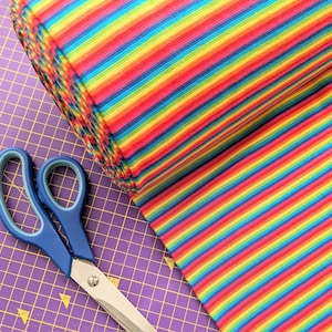 Könnte beinhalten: Eine Rolle regenbogenfarbener gestreifter Stoff auf einer lila Schneidematte mit einer blauen Schere.