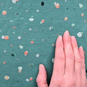 Op de afbeelding: Een teal stof met een gespikkeld patroon van witte, oranje en zwarte stippen. Een hand rust op de stof.