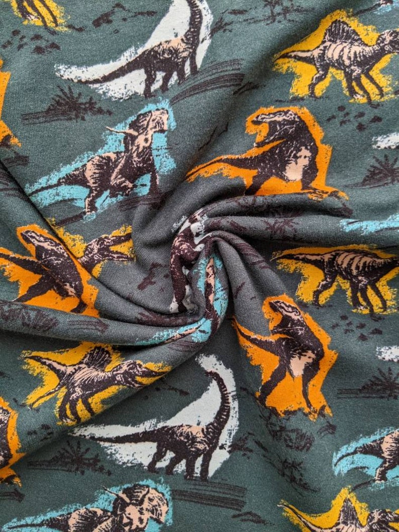 May include: Green fabric with a repeating pattern of dinosaurs in various colors, including orange, yellow, white, and black.