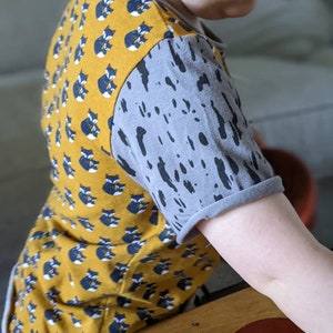 K&ouml;nnte beinhalten: Ein gelbes und graues T-Shirt mit kurzen &Auml;rmeln, das auf der Vorderseite ein Muster aus schwarzen und wei&szlig;en F&uuml;chsen und auf den &Auml;rmeln ein graues Spritzmuster hat.