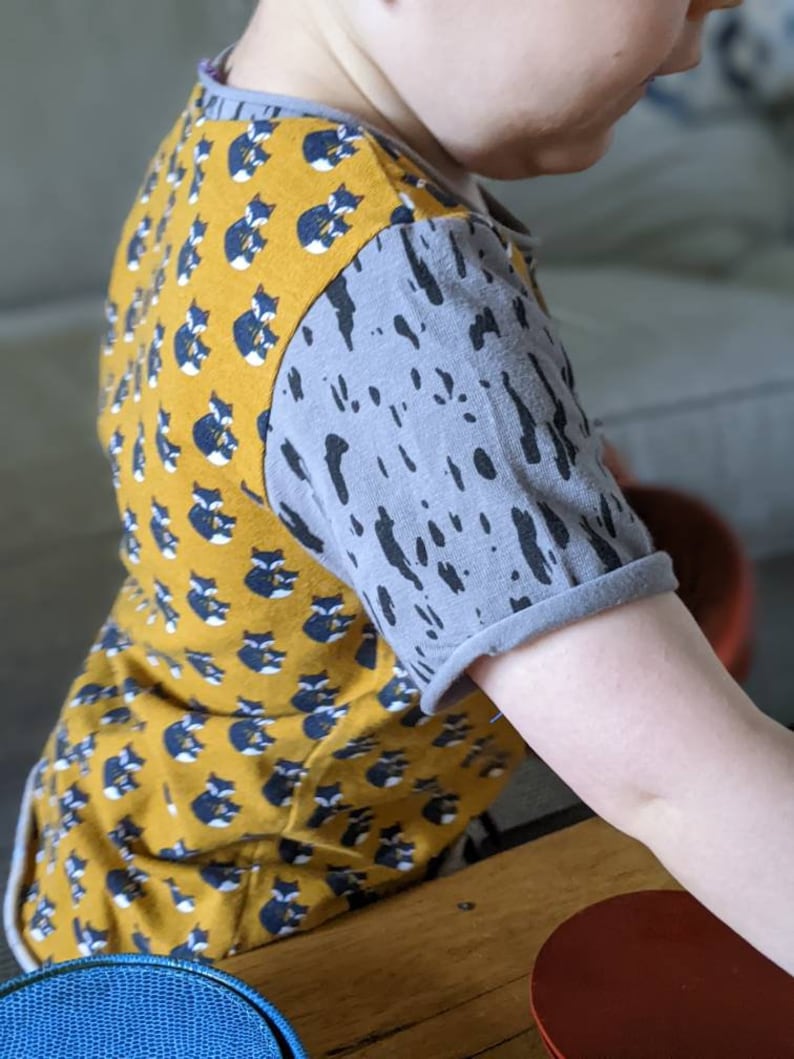 K&ouml;nnte beinhalten: Ein gelbes und graues T-Shirt mit kurzen &Auml;rmeln, das auf der Vorderseite ein Muster aus schwarzen und wei&szlig;en F&uuml;chsen und auf den &Auml;rmeln ein graues Spritzmuster hat.