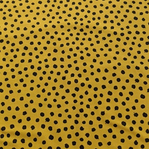 Op de afbeelding: Gele stof met een zwart stippenpatroon. De stippen zijn enigszins onregelmatig in vorm en grootte.