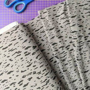 K&ouml;nnte beinhalten: Grauer Stoff mit einem schwarzen abstrakten Druck. Der Stoff ist gefaltet und eine blaue Schere liegt auf einer violetten Schneidematte.