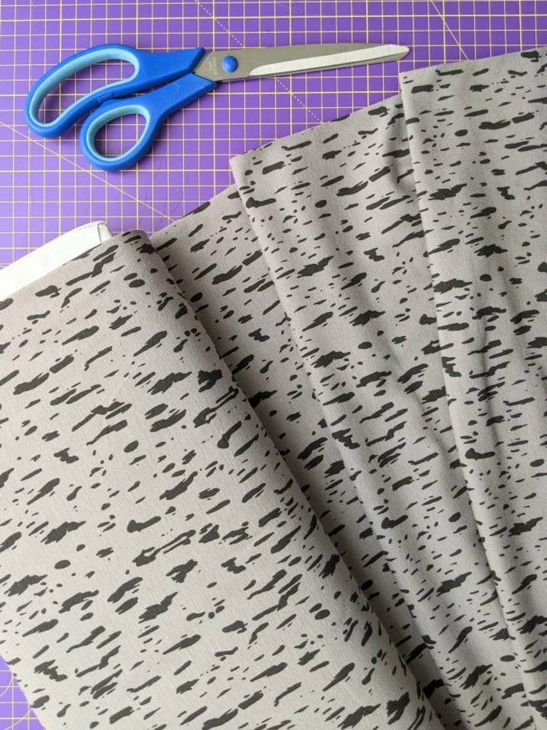 K&ouml;nnte beinhalten: Grauer Stoff mit einem schwarzen abstrakten Druck. Der Stoff ist gefaltet und eine blaue Schere liegt auf einer violetten Schneidematte.