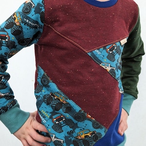 K&ouml;nnte beinhalten: Ein lang&auml;rmliges Shirt f&uuml;r Kinder mit Patchwork-Design. Das Shirt besteht aus verschiedenen Stoffen in einem blau-gr&uuml;n-roten Farbschema. Der Stoff hat einen Monstertruck-Druck mit den Worten "Vroom!" und "Speed!" darauf.
