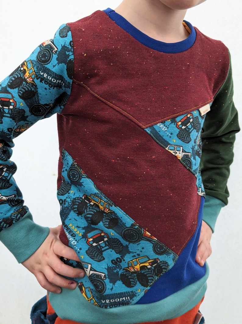 K&ouml;nnte beinhalten: Ein lang&auml;rmliges Shirt f&uuml;r Kinder mit Patchwork-Design. Das Shirt besteht aus verschiedenen Stoffen in einem blau-gr&uuml;n-roten Farbschema. Der Stoff hat einen Monstertruck-Druck mit den Worten "Vroom!" und "Speed!" darauf.
