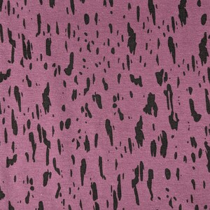 May include: A close-up view of a mauve fabric featuring a pattern of black, irregular shapes, reminiscent of paint splatters. The shapes vary in size and density, creating a textured effect. The fabric appears to be a knit or jersey material.