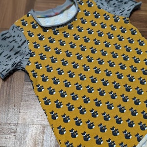 K&ouml;nnte beinhalten: Ein gelbes T-Shirt mit kurzen &Auml;rmeln und einem schwarzen und wei&szlig;en Fuchsmuster. Das Shirt hat einen grau-wei&szlig;en gemusterten Stoff an den &Auml;rmeln.