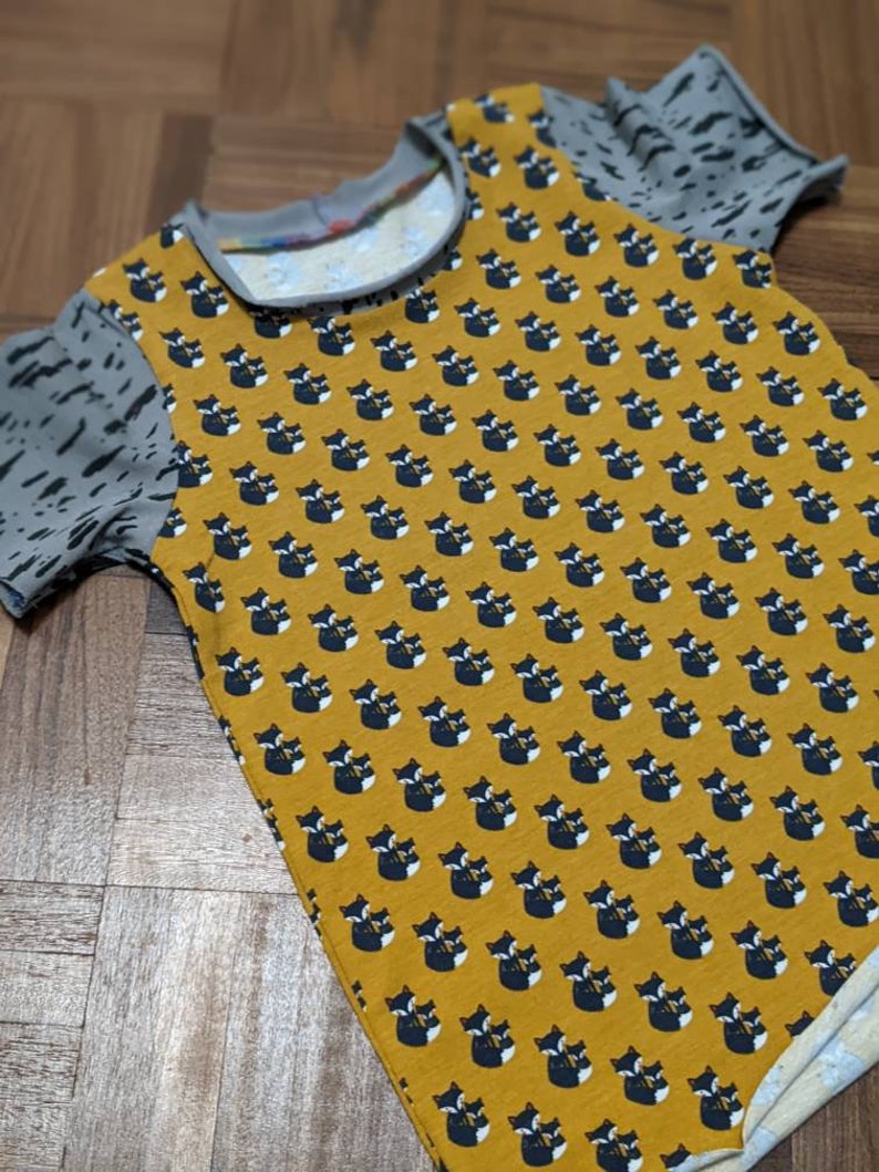K&ouml;nnte beinhalten: Ein gelbes T-Shirt mit kurzen &Auml;rmeln und einem schwarzen und wei&szlig;en Fuchsmuster. Das Shirt hat einen grau-wei&szlig;en gemusterten Stoff an den &Auml;rmeln.